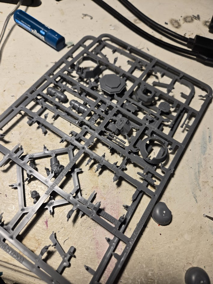 Gray plastic model‑kit sprue laid out on a hobby work surface, showing unassembled miniature parts alongside scattered tools and paint marks.